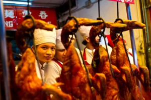 Young man in white hat and apron behind a row of hanging barbecued ducks