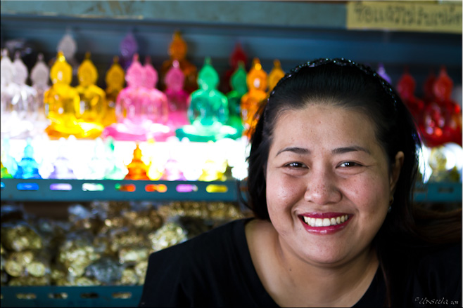 Portrait: Smiling Thai woman in front of shelves of colourful buddhas