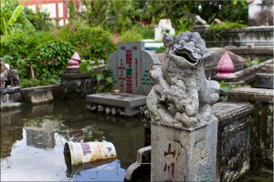 A Chinese stone lion in front of a gravesite filled with water and rubbish.