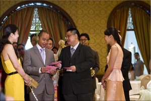 Two young Thai women hold an rope made of flowers, as two Thai men negotiate red envelopes.