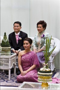Family portrait: a young Thai woman in formal attire seated on the floor in front of a middle aged Thai couple seated on a sofa.