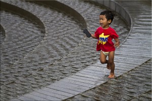 Young child in a red t-shirt and diaper running through a water fountain.