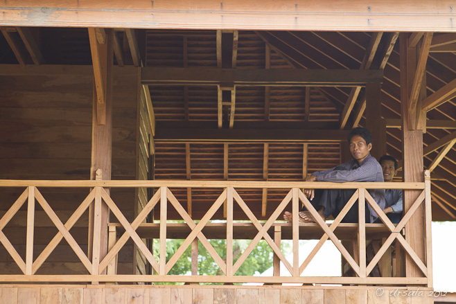 A khmer man sitting on timber framing on the balcony of an unfinished wooden building.