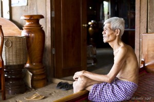 Portrait: Gray-haired elderly Khmer man seated on a wooden bench in his purple checkered woven cotton 