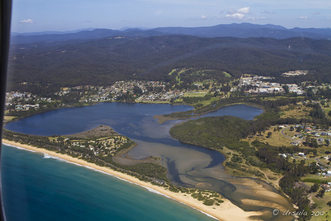 Aerial view: green sea waters, long white beach, dark blue estuary, blue-green hills and a blue sky.