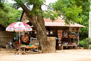 Khmer man under a large umbrella cutting guava in front of a rural mixed-goods store, Cambodia.