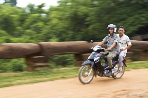 Two Khmer men on a small motorcycle crossing an old Angkor bridge