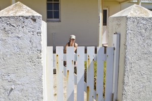 Young woman in a cap sits in the sun behind a white stone wall and picket gate.