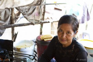 Portrait: Khmer lady in a market, a bucket of batter and some cooked waffles in the background.