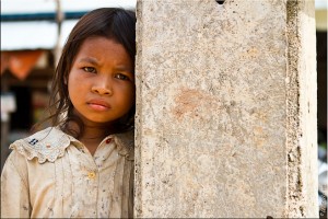 Portrait: Sad-faced khmer girl in a dirty white blouse resting against a cement pillar