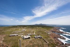Scene: view down over three homesteads on flat land; mountains and blue skies in the distance.