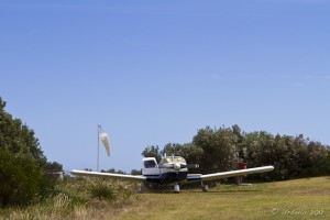 A Cherokee 6 on a grass runway, an empty wind sock behind.