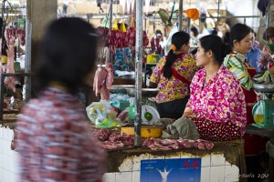 Scene: Busy meat market in Cambodia: sellers sitting on tables, shoppers passing