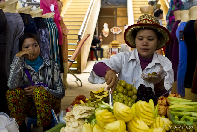 Khmer woman in a straw hat selling cut fruit as another woman looks on.