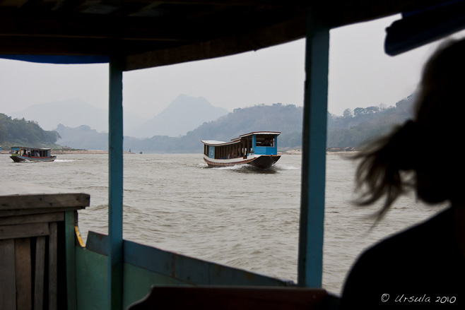 View of a traditional wooden Lao river boat, from another. Mekong River and hills in the background.