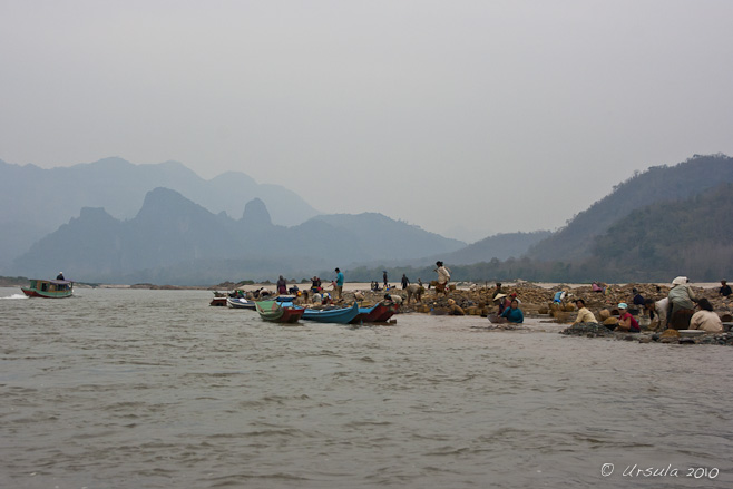 View of people and long wooden boats on sandbars on the river: ragged karst mountains in the background.