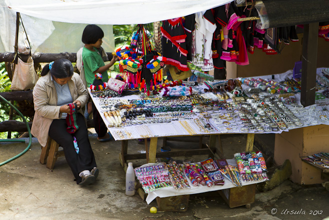 Akha woman sits embroidering next to a table of market wares.