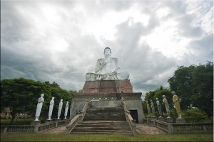 View: Large seated Buddha flanked by standing statues, cloudy sky.
