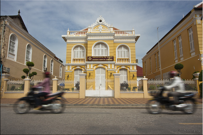 Two scooters blurring past a yellow French colonial building.