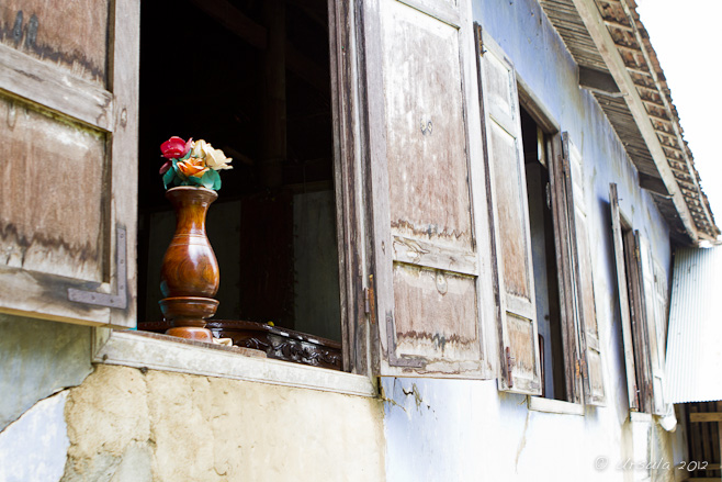 Artificial flowers in a wooden vase in and open window with wooden shutters.