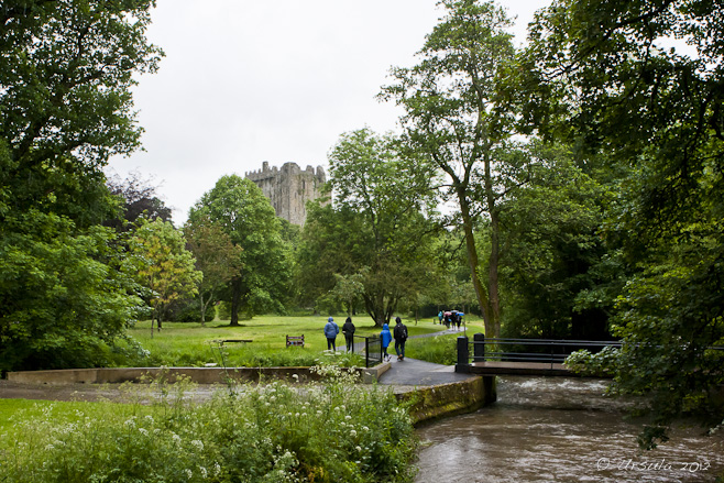 Small bridge over a muddy river - green lawns, trees and Blarney Castle in the background.