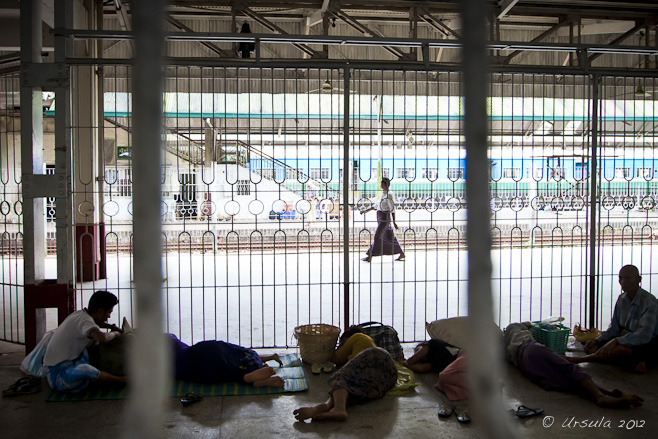 Man in a longyi on a bright railway platform, behind the rails of the waiting room.