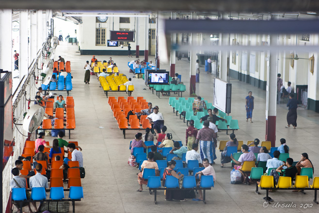 A scattering of people in banks of colourful plastic seats in a railway waiting area.