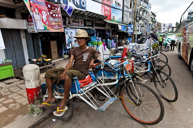 A burmese bicycle rickshaw driver sits on his vehicle in a busy market street. Bago, Myanmar