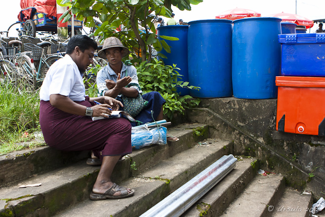 Two Burmese men sitting on cement steps; one telling a story, the other writing.