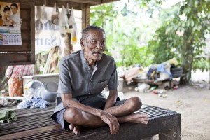 Elderly Thai man sitting cross-legged on a wooden platform.