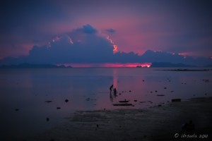Dark blue and pink sky over calm sea with two people silhouetted.