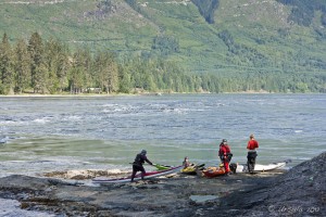 Kayakers in helmets and wet-weather gear stand on the rock at Roland Point, Skookumchuck