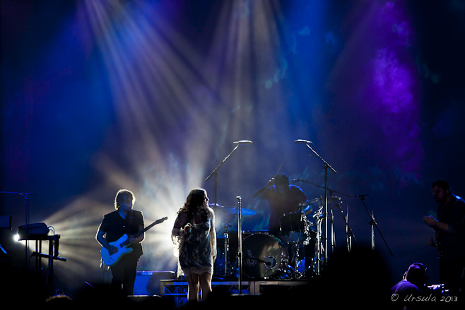 Dark blue stage with singer and band backlit by white light.