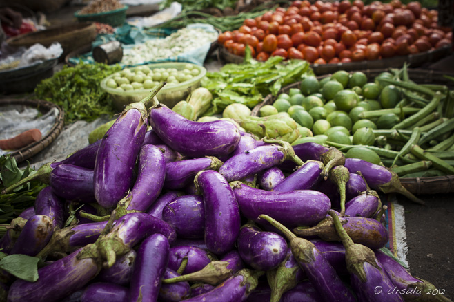 Purple eggplant, green limes, red tomatoes and other vegetables in baskets on the ground in an outdoor Mandalay street market.