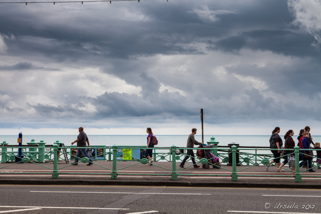 Warmly-dressed people on and ocean-front walkway, under black skies, Kings Rd Brighton, UK