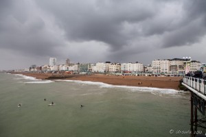 View from Brighton Pier back to the beach and low-rise buildings behind; body boarders in the water under low black clouds.