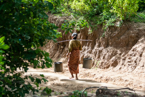Burmese woman walking away on a dirt path with water buckets on a yoke, Myanmar