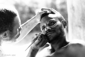 B&W portrait of a Papuan man being face-painted with cassowary eyes, Middle Sepik, PNG