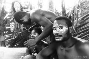 Environmental B&W portrait of a Papuan man being face-painted with cassowary eyes, Middle Sepik, PNG