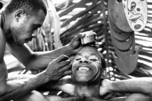 B&W portrait of a Papuan man being face-painted with cassowary eyes, Middle Sepik, PNG