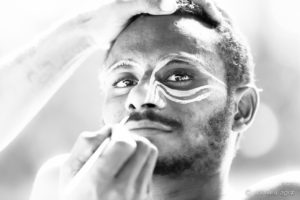 B&W portrait of a Papuan man being face-painted with cassowary eyes, Middle Sepik, PNG