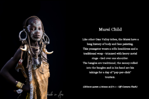Picture: Mursi child in a nilla headdress and a traditional wrap tied over one shoulder. Text: the money rolled into the bangles and in his hand are his takings for a day of “pay-per-click” tourism.
