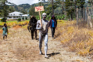 Clay-painted man with a signboad reading: Omo Masalai from Simbu, Mt Hagen Papua New Guinea