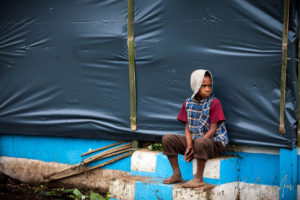 Seated Papuan child on a steps in front of a plastic-sheeted building, Mt Hagen PNG.